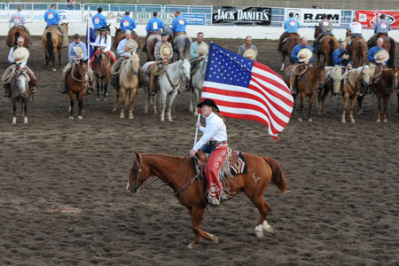 ©Bill Patterson, Patterson Photography photo showing a cowboy on a horse holding an American flag riding in front of mounted cowboys holding their hats in a rodeo ring