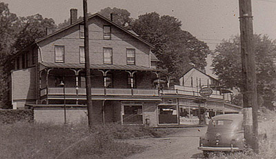a 1940s sepia photograph of a car parked across a dirt road across the railroad tracks from a 4 story converted hotel with a store on the lowest level and a balcony above the store